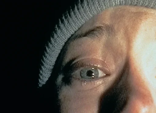 A shadowy figure in a dark forest, reminiscent of The Blair Witch Project's eerie aesthetic.