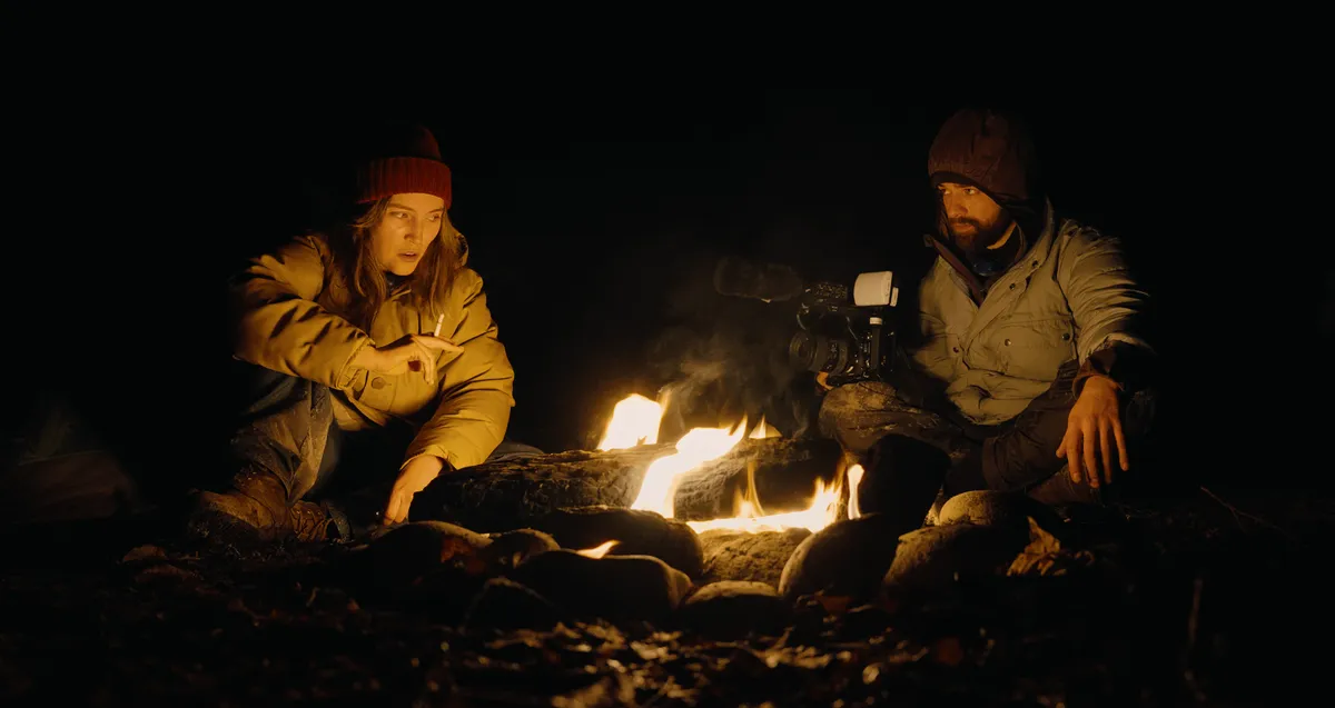 A dark, grainy still from a found-footage horror movie, showing a dimly lit, possibly wooded area, suggesting mystery and fear.