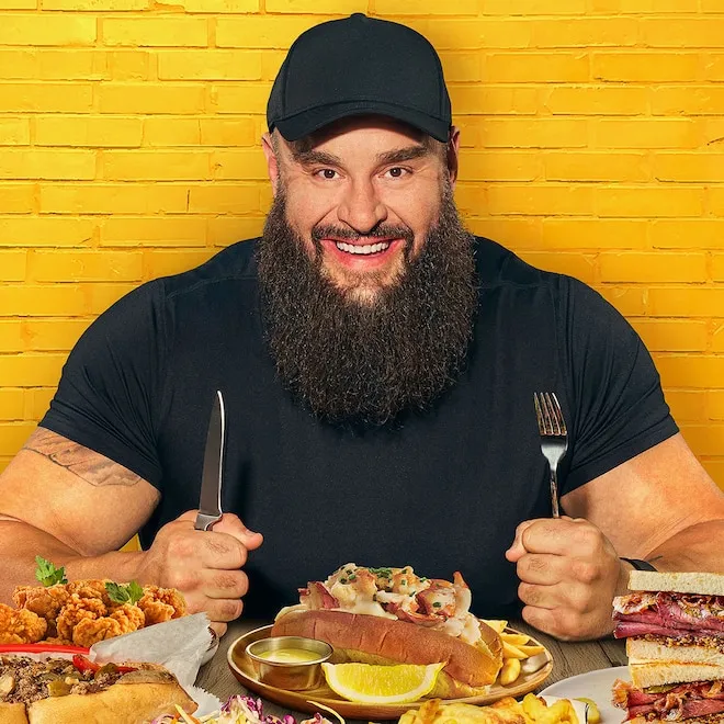 Braun Strowman, a muscular WWE wrestler, smiling while holding a plate of food.