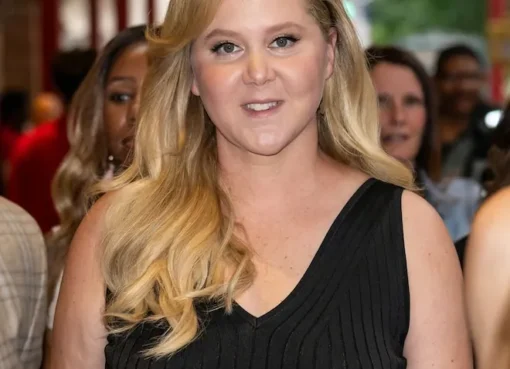 Amy Schumer smiling in a bright, airy living room with large windows.