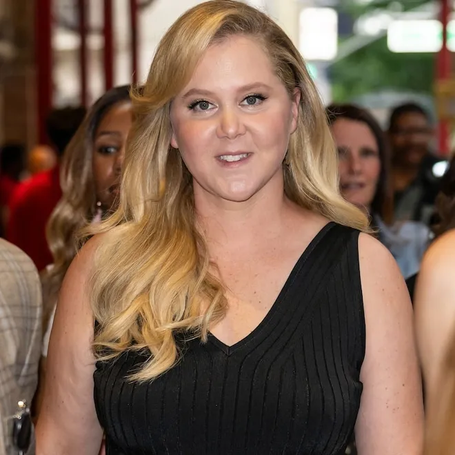Amy Schumer smiling in a bright, airy living room with large windows.