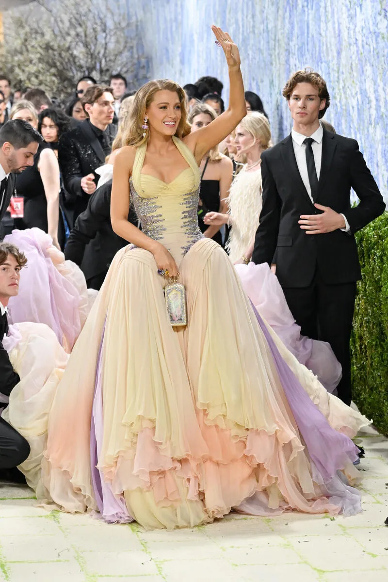 Blake Lively smiling and posing on the Met Gala red carpet in a beautiful multi-colored, layered pastel gown.
