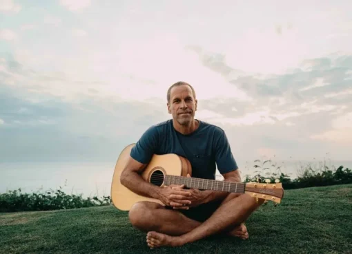 Jack Johnson standing by the ocean with a surfboard, looking relaxed.