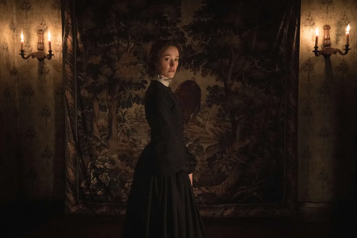 Maika Monroe as a crazed governess, with a wide unsettling smile, in a dark, gothic Victorian setting.