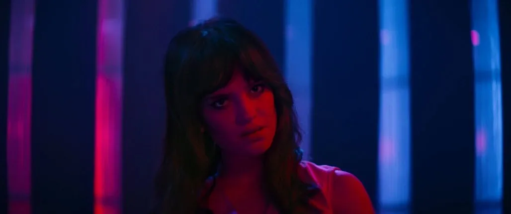 A dark, moody still from a film, with neon lights reflecting on a character's face, hinting at a surreal, unsettling atmosphere.