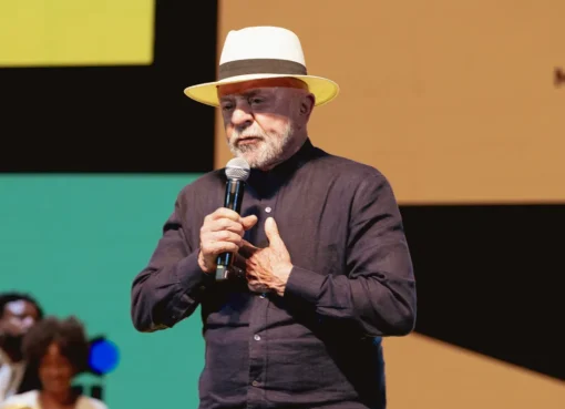 President Lula smiling and speaking at an event, with a screen displaying the 'Tela Brasil' logo and graphics.