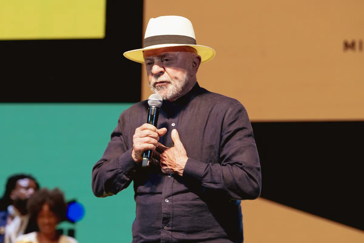 President Lula smiling and speaking at an event, with a screen displaying the 'Tela Brasil' logo and graphics.