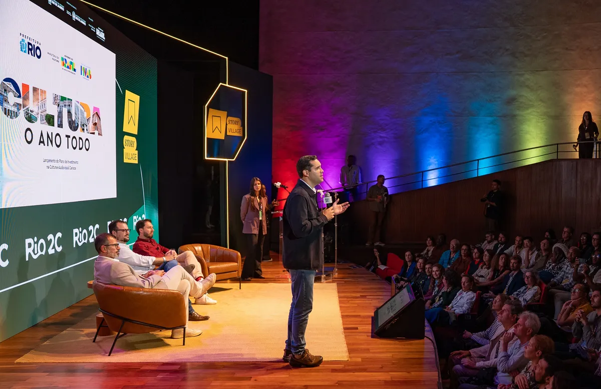 Rio de Janeiro Mayor Eduardo Cavaliere smiling and speaking at a creative industry event, possibly Rio2C, with a backdrop of colorful art or digital screens.