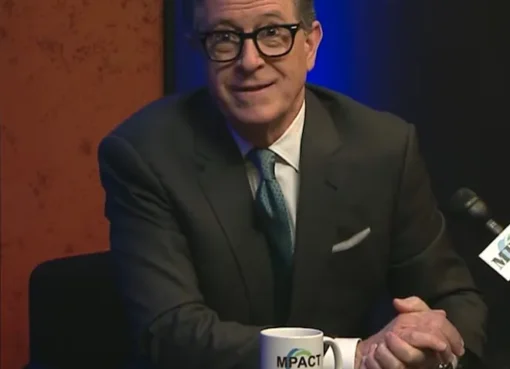 Stephen Colbert smiling cheerfully on a television screen.