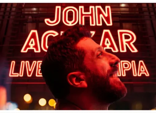 John Achkar performing stand-up comedy on a stage at L'Olympia Hall.