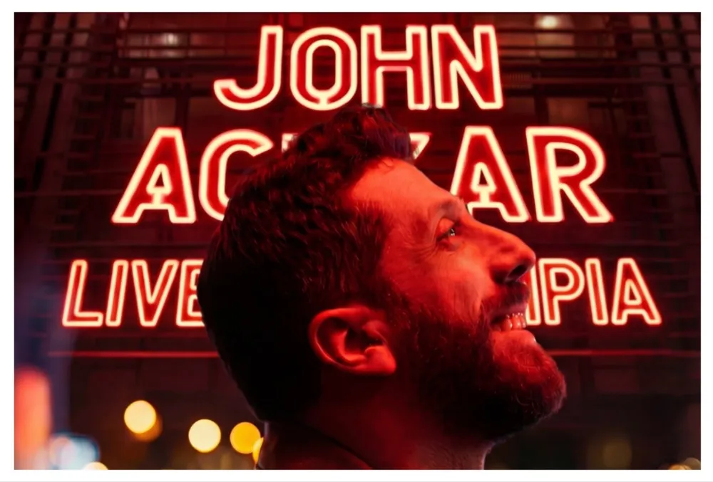John Achkar performing stand-up comedy on a stage at L'Olympia Hall.