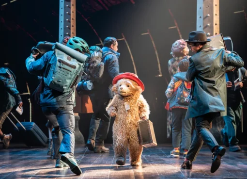 Paddington Bear wearing his signature blue coat and red hat, standing on a Broadway stage with bright lights.