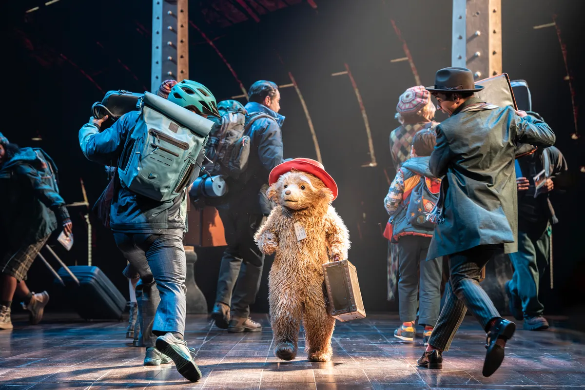 Paddington Bear wearing his signature blue coat and red hat, standing on a Broadway stage with bright lights.