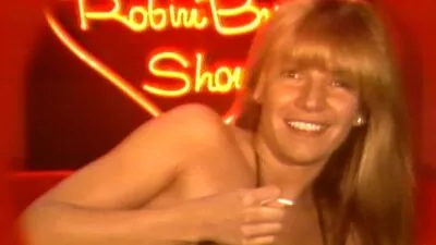 Robin Byrd smiling brightly, wearing a sequined top and big hair on a retro television set.