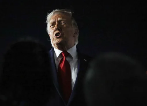 Donald Trump speaking at a podium, with flags in the background, looking determined.