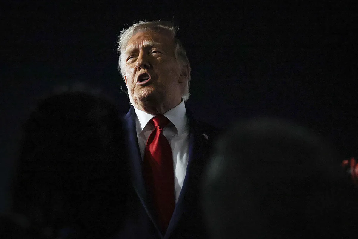 Donald Trump speaking at a podium, with flags in the background, looking determined.