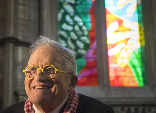 A photograph of David Hockney, smiling, possibly next to one of his colorful artworks.