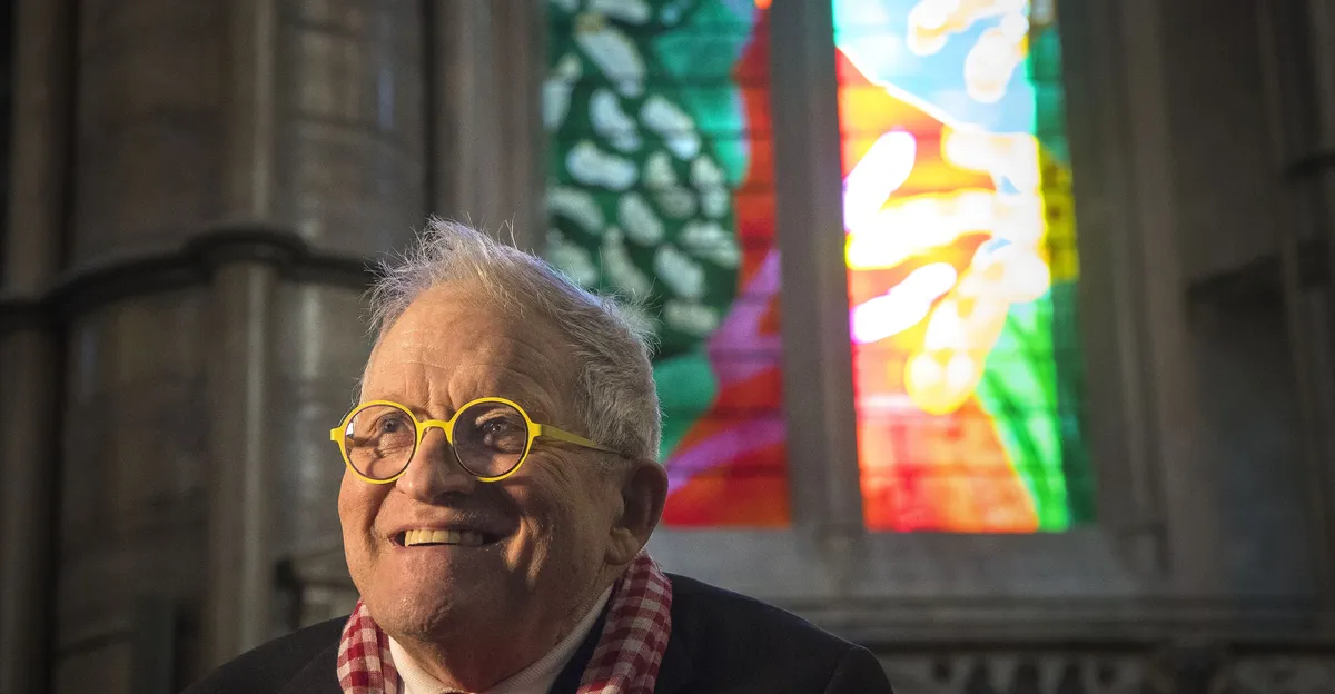 A photograph of David Hockney, smiling, possibly next to one of his colorful artworks.