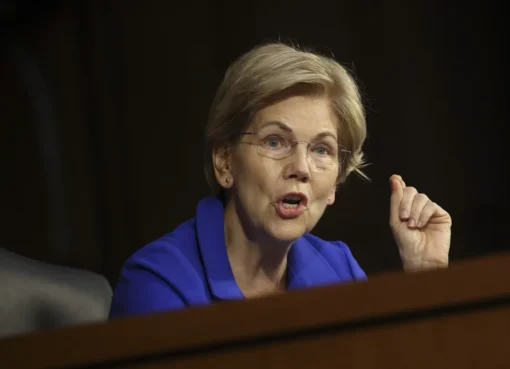 Senator Elizabeth Warren speaking seriously at a podium with a subtle blue and white background.