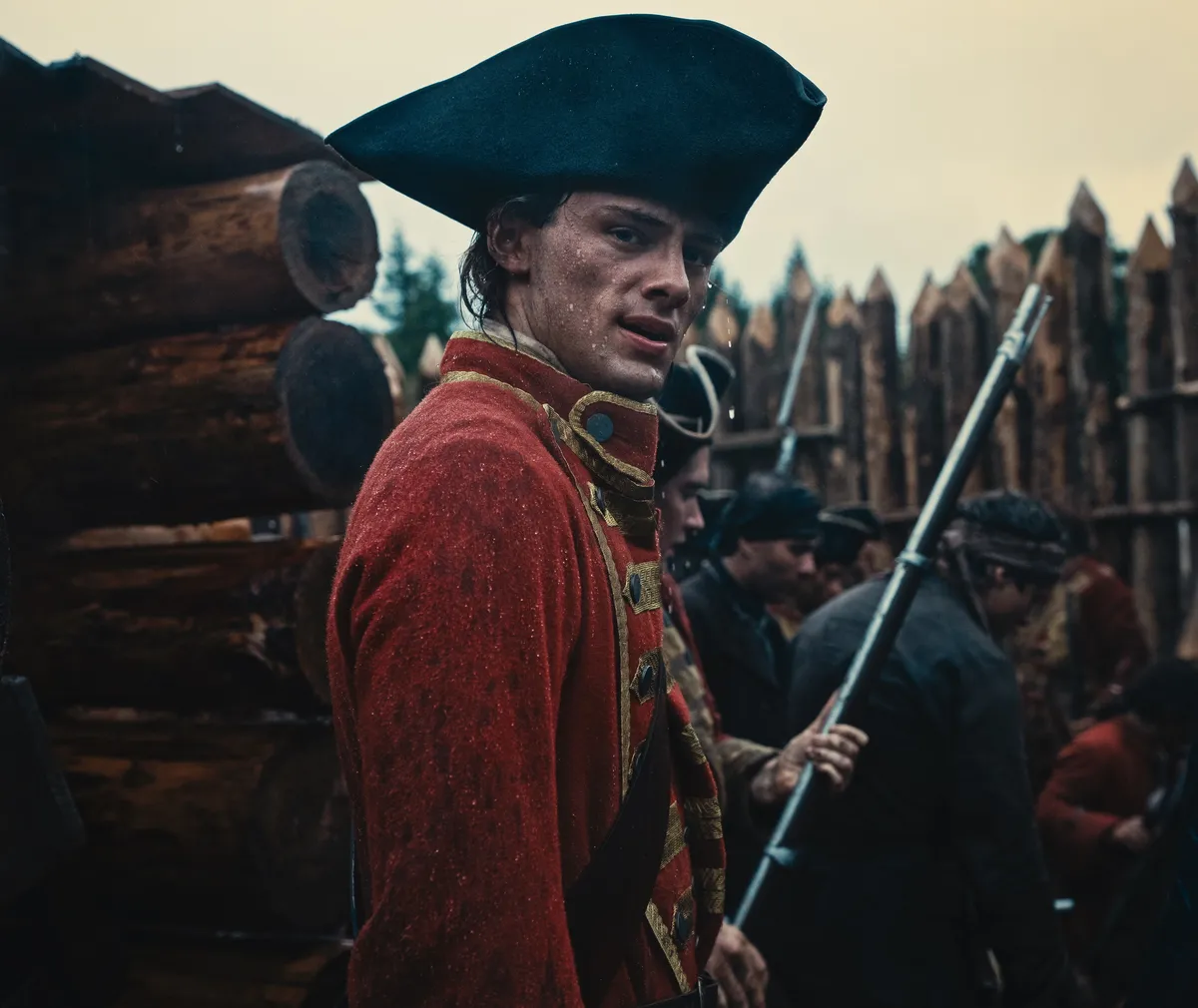 A young man in 1700s clothing standing in a sunlit forest looking determined.