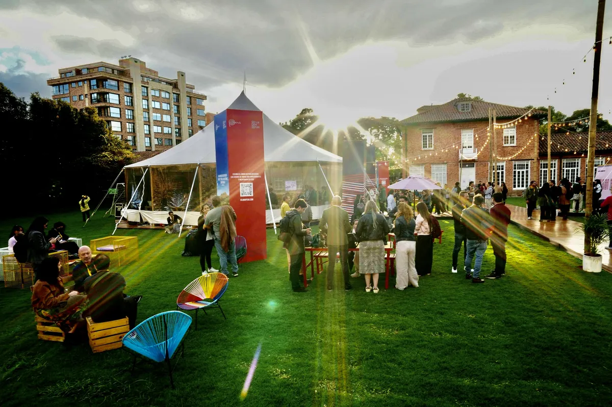 A group of diverse young filmmakers networking at the Bogotá Audiovisual Market, with film equipment and colorful banners in the background.