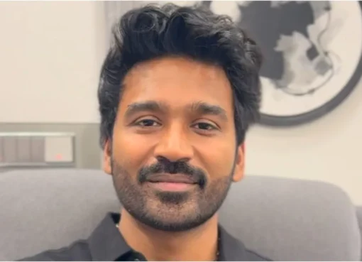 Dhanush smiling, possibly in front of a luxury home or car, representing his wealth.