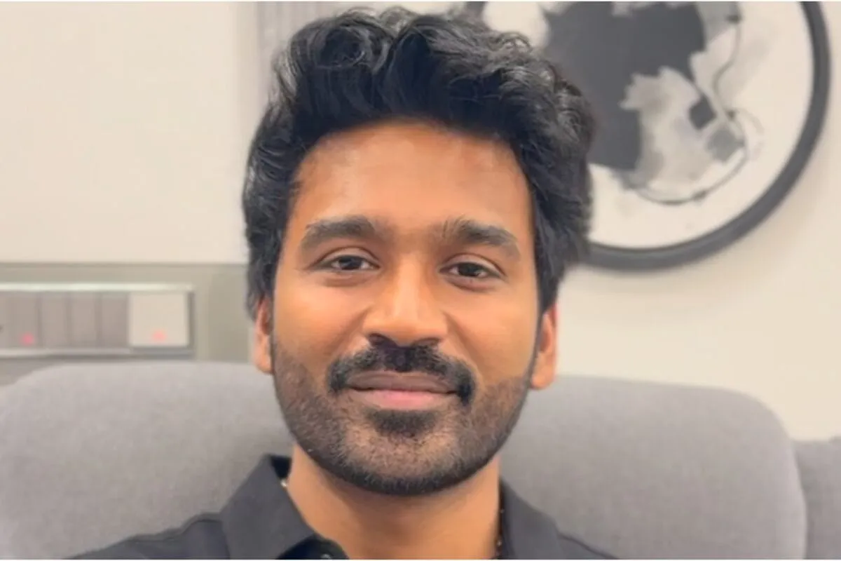 Dhanush smiling, possibly in front of a luxury home or car, representing his wealth.