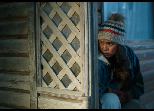 A young Métis woman named Elyse looks over her shoulder with a concerned expression in a dark, atmospheric forest setting, hinting at a hidden threat.