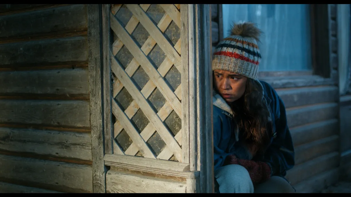 A young Métis woman named Elyse looks over her shoulder with a concerned expression in a dark, atmospheric forest setting, hinting at a hidden threat.
