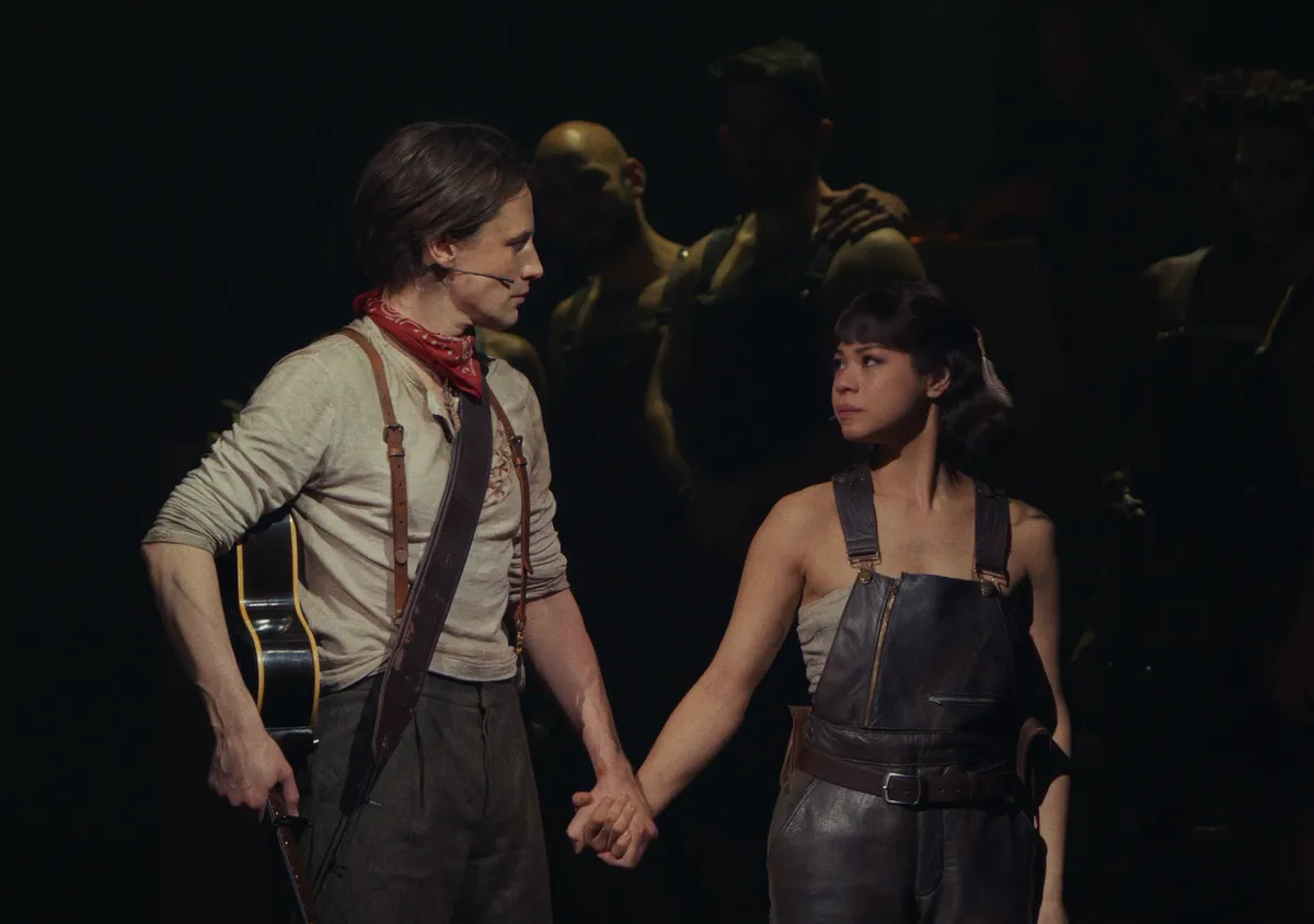 A stylized image of characters from Hadestown, possibly Hades and Persephone, in a moody, jazz-club-like underworld setting with warm and cool lighting.