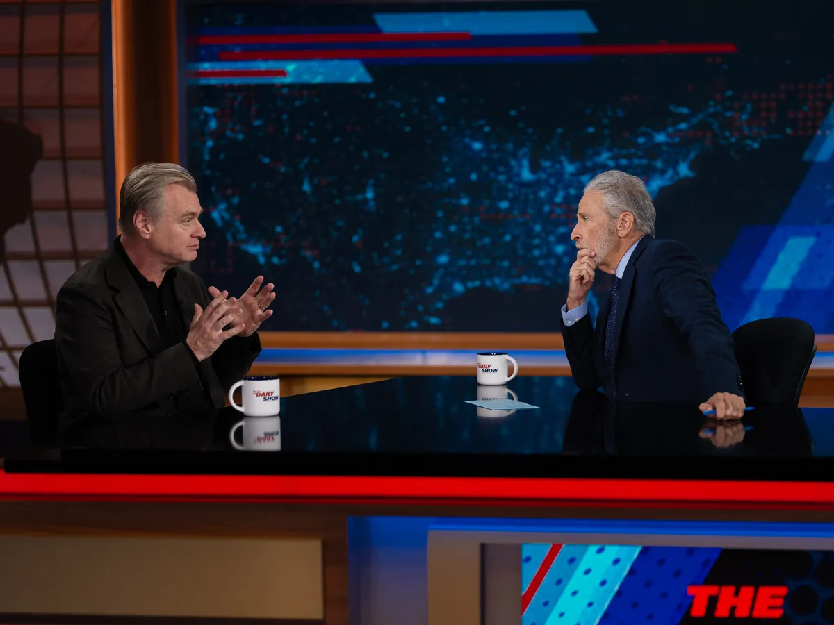 Christopher Nolan and Jon Stewart sharing a laugh on The Daily Show set.