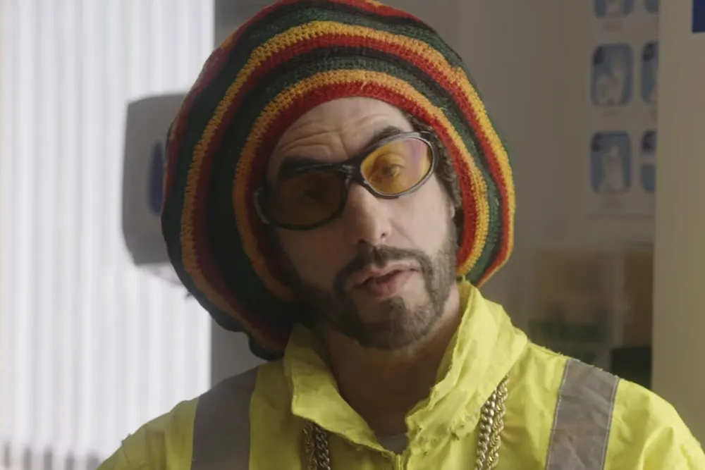 Ali G in his signature yellow tracksuit and beanie, looking surprised or defiant.