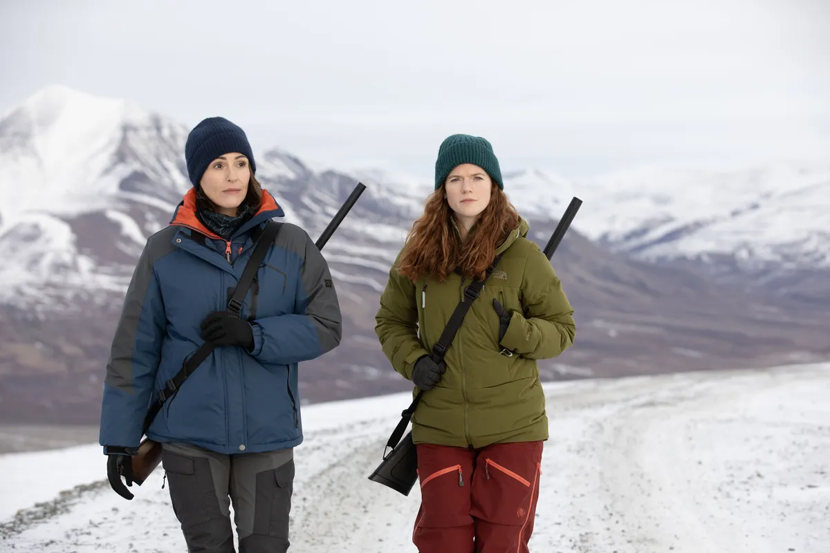 Suranne Jones and Rose Leslie in winter gear, trekking across a snowy Arctic landscape with rifles.