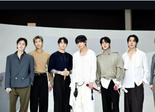 BTS members smiling and waving on stage under bright lights.