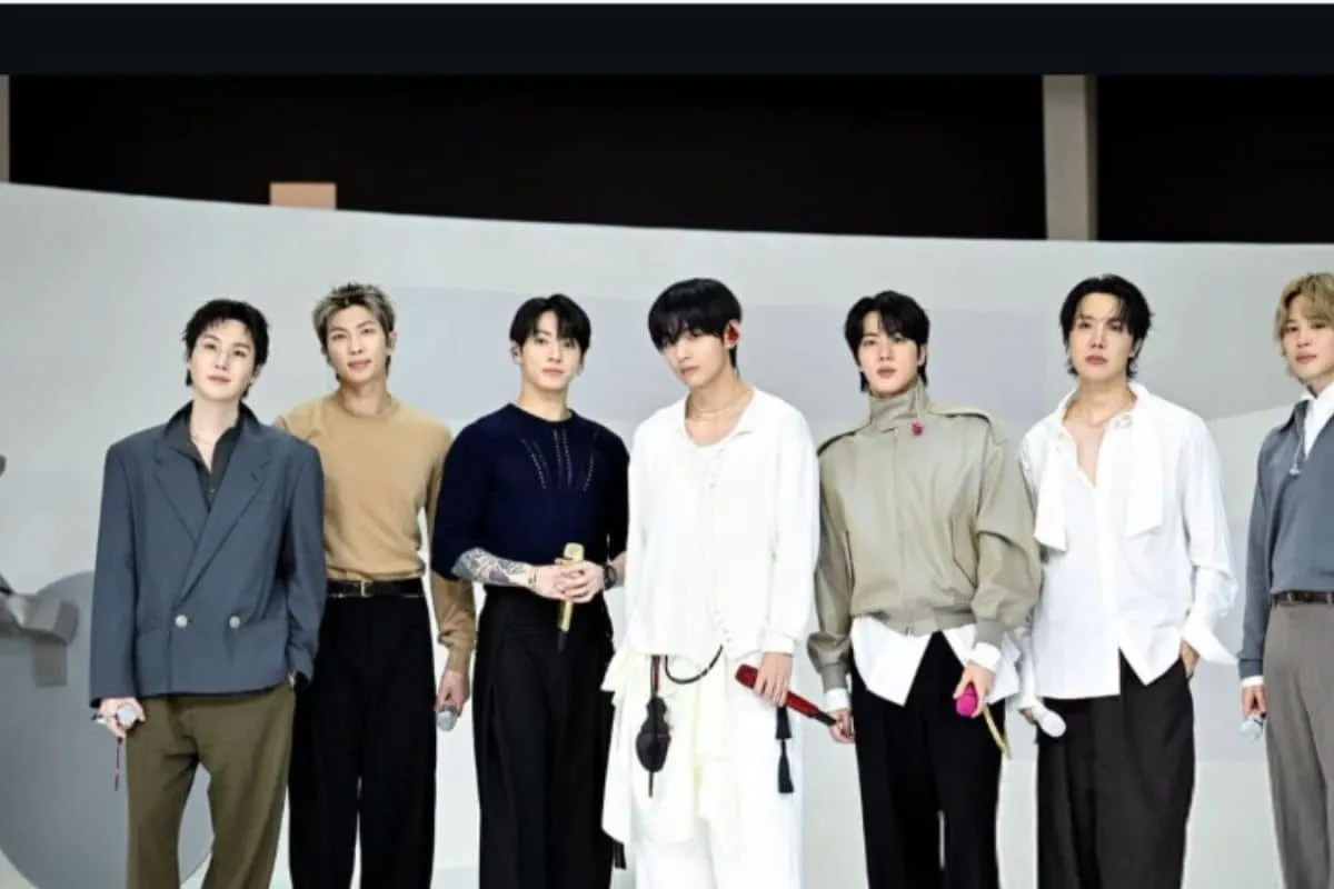 BTS members smiling and waving on stage under bright lights.