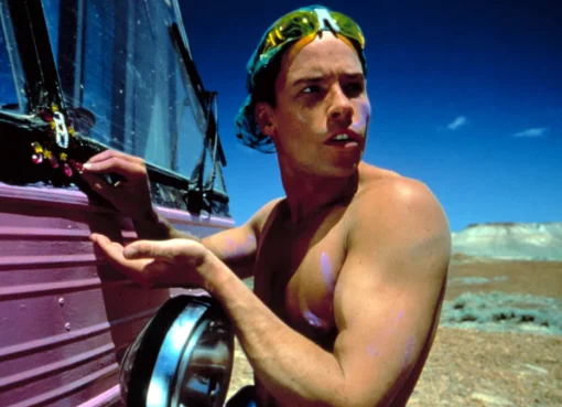 Guy Pearce in a bright, colorful costume from Priscilla, Queen of the Desert, standing in the Australian Outback.