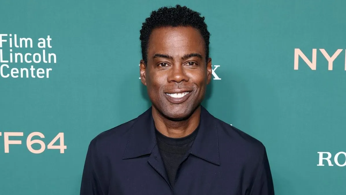 Chris Rock smiling, looking thoughtful, with a microphone nearby.