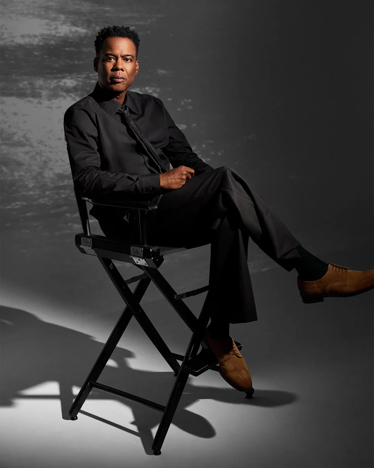 Chris Rock looking thoughtful, possibly on a film set, with a slight smile.