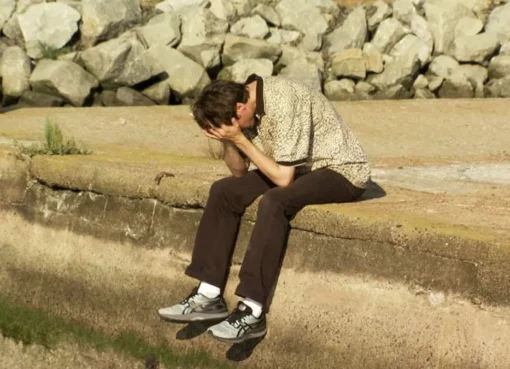 A comedian looking thoughtfully at old video footage, perhaps from a camcorder screen.