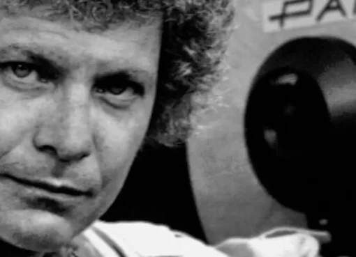 A black and white photo of a smiling David Worth holding a film camera on a movie set.