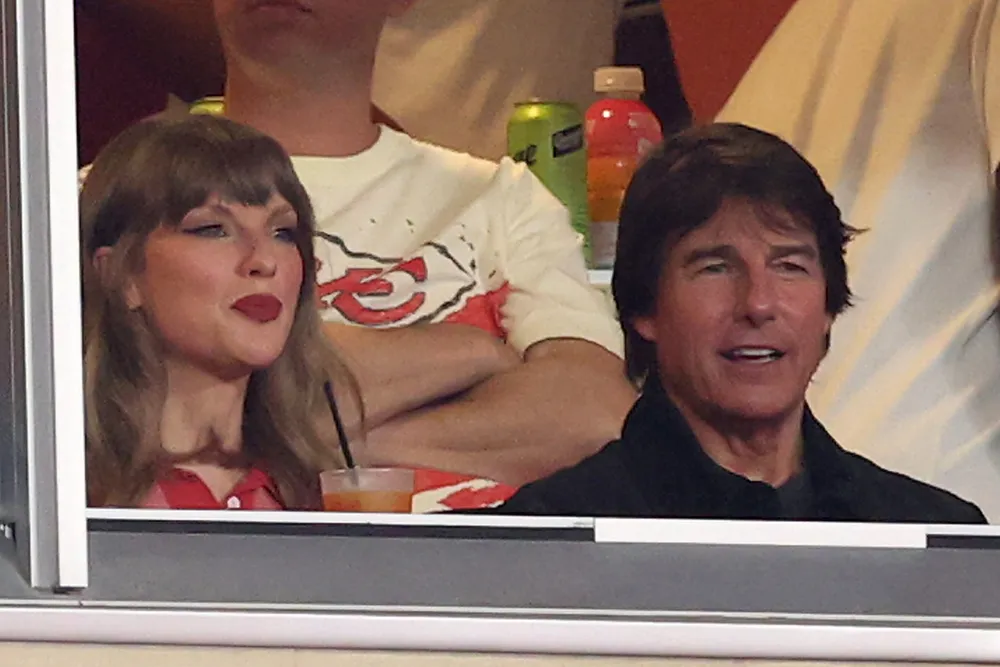 Tom Cruise and Taylor Swift sitting side-by-side at a football game, smiling and talking.