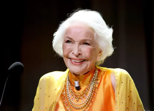 Ellen Burstyn, a senior woman with short white hair, smiling and holding a golden lion award, possibly wiping a tear from her eye on stage.