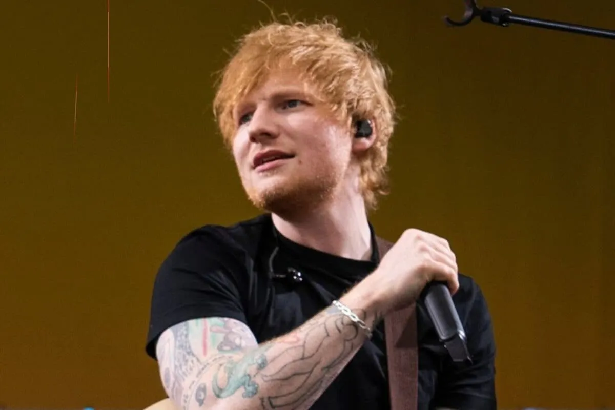 Ed Sheeran on stage with a guitar, a spotlight shining on him, at a concert.