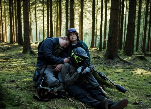 Three unclothed teenagers stand close in a forest, gently caressing wounds on each other.