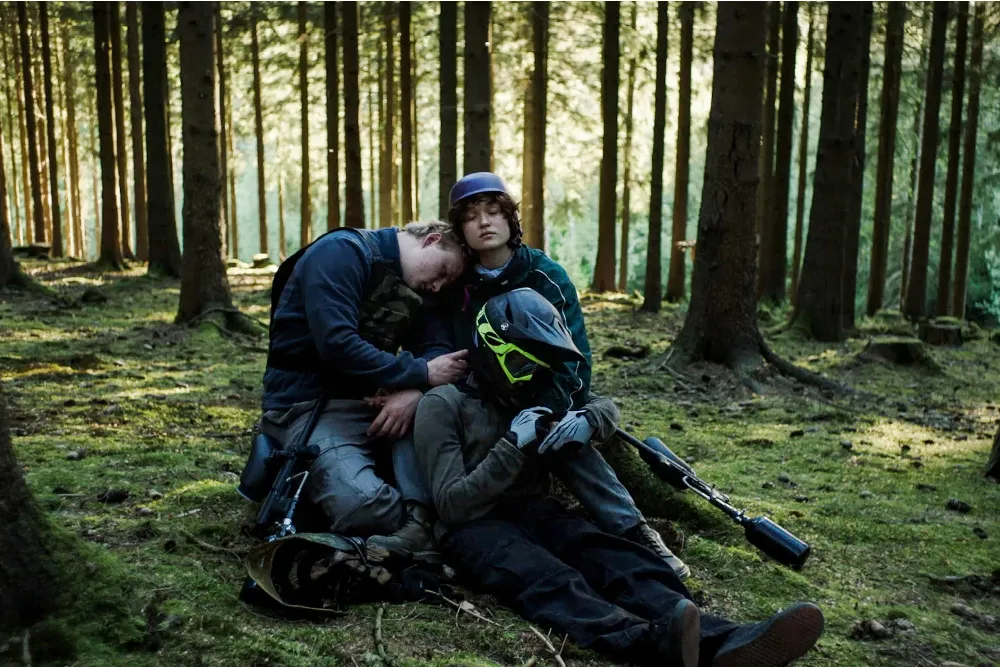 Three unclothed teenagers stand close in a forest, gently caressing wounds on each other.
