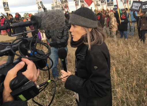 A determined woman, Amy Goodman, speaking into a microphone with a serious expression.