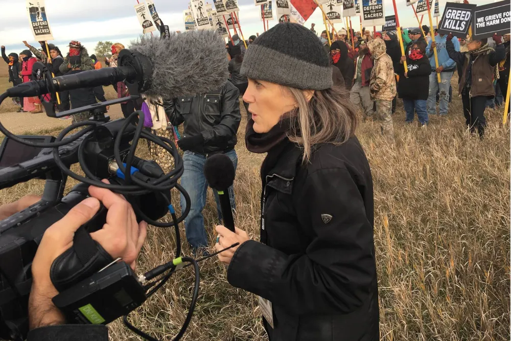 A determined woman, Amy Goodman, speaking into a microphone with a serious expression.