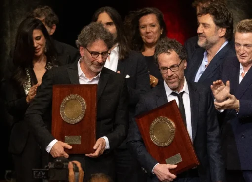 Joel and Ethan Coen accepting an award on stage, smiling, with a crowd in the background.