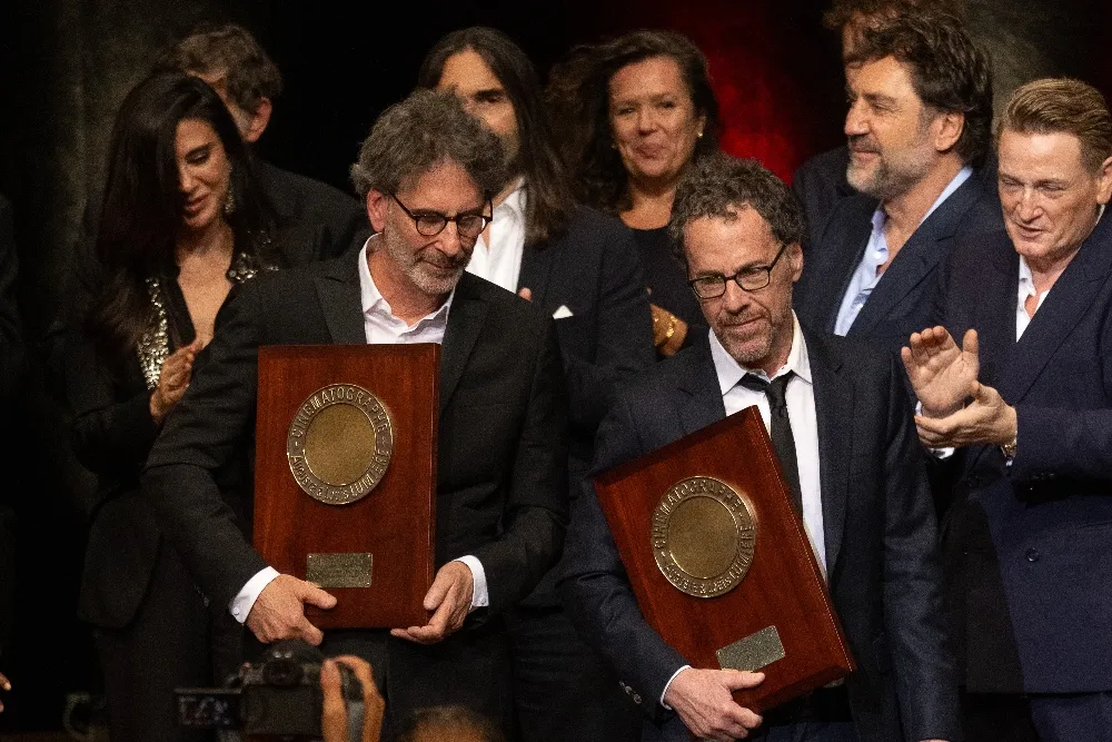 Joel and Ethan Coen accepting an award on stage, smiling, with a crowd in the background.