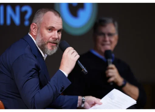 A man with a thoughtful expression speaking at a conference, the room has a modern, clean aesthetic with cool blues and muted grays.